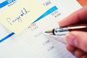 A fountain pen checks a financial or business document with an adhesive note reading "payroll" attached to it. 