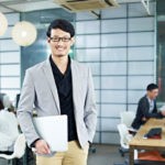 Picture of young asian entrepreneur standing in office with laptop computer under arm.